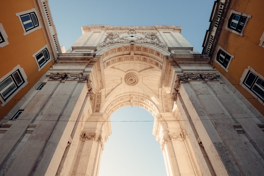 Looking from below, up above to the  Rua Augusta Arch in Lisbon, with white marble archway and yellow buildings around it.