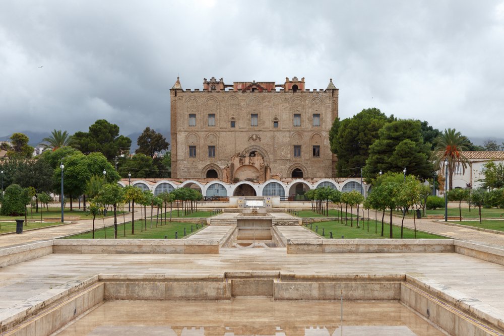 A small brown-toned castle with a garden in front of it on a cloudy day in Palermo.