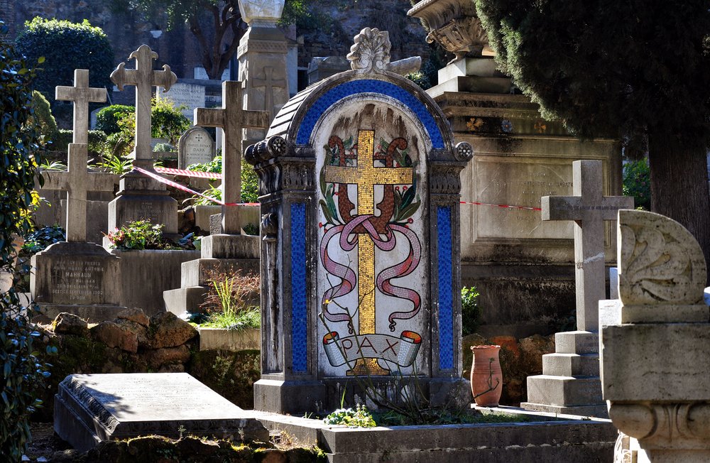 Protestant Cemetery in Rome with a colorful mosaic-tiled headstone with blue, gold, red detailing and the word 'pax' which is latin for peace