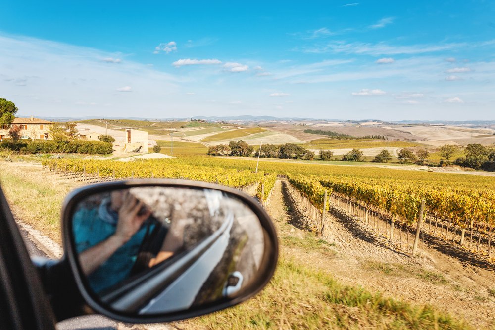 View from car to the Tuscany wine fields, you can see the rearview mirror and someone taking a photo in the mirror.