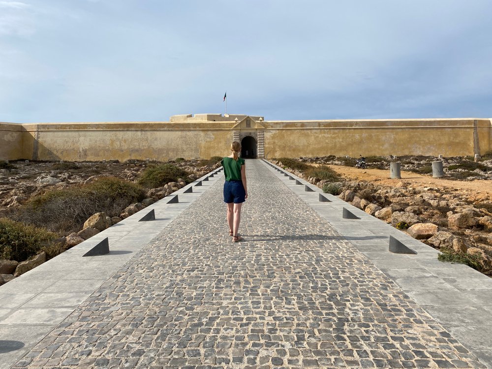 Tourist woman on paved road to entrance of historic Sagres Fortress (Fortaleza de Sagres), old landmark ruin attraction in the Algarve region of Portugal
