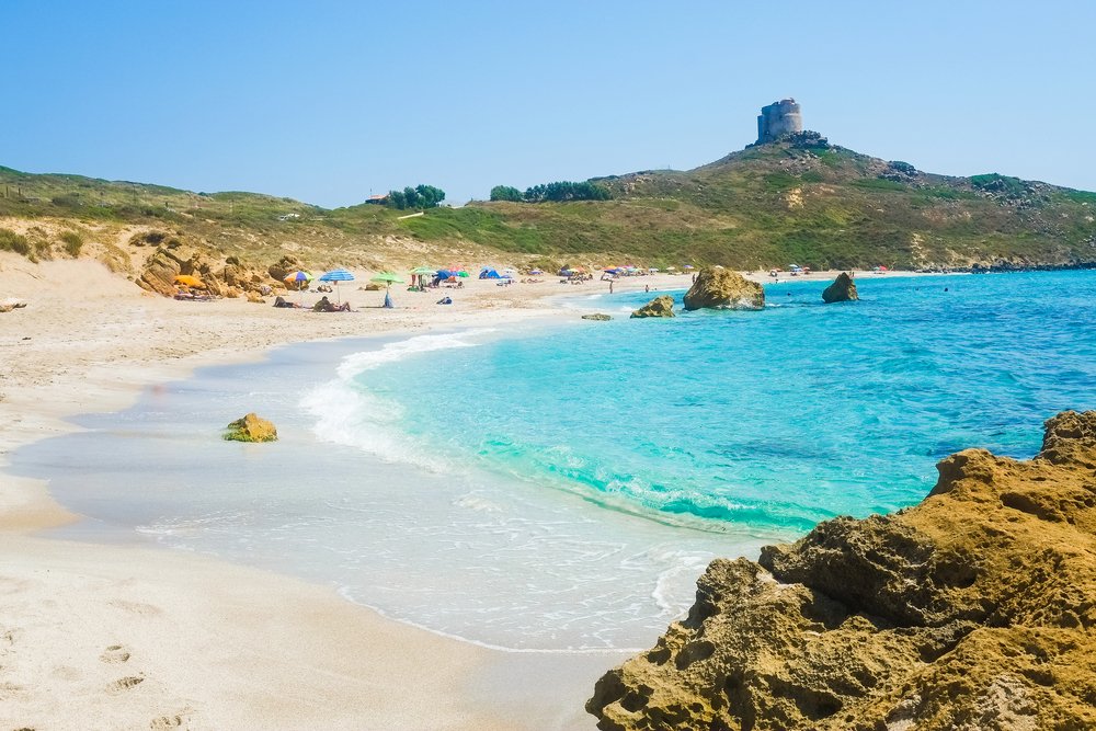 San Giovanni di Sinis beach in Sardinia, with a beach with people with umbrellas lounging in the sun, and brilliant turquoise waters and ruins in the distance.