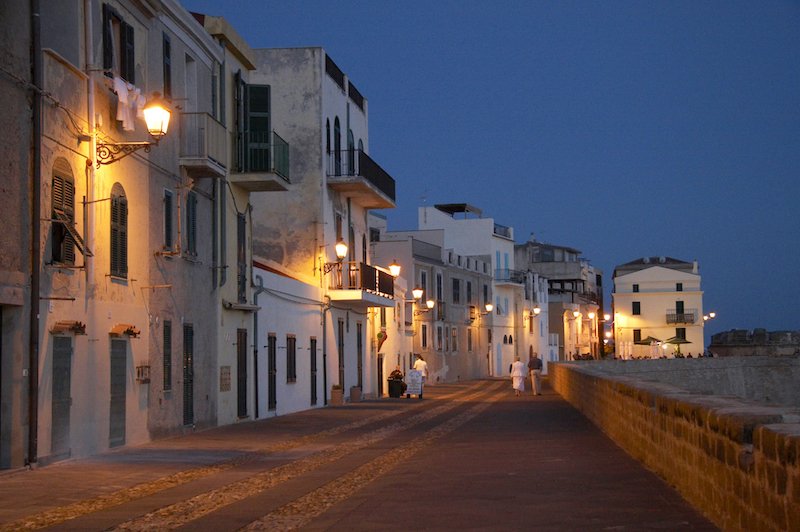 A nighttime stroll in the waterfront area of Alghero, with some lights on, and just a few people on the street.