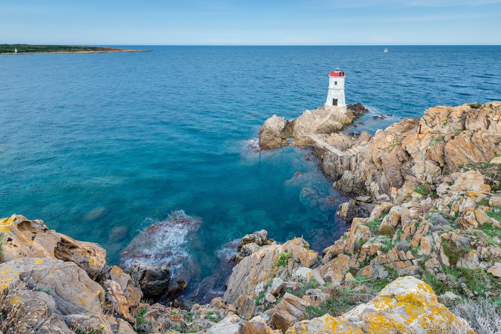 Capo Ferro cape lighthouse in Sardinia, Italy, on the edge of a cliff with stairs leading down to it, surrounded by clear blue water