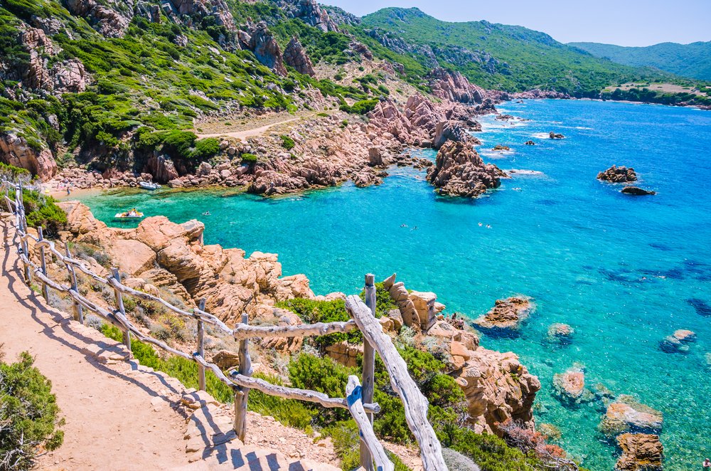 Stony walk path in Costa Paradiso in Sardinia, Italy, with a wooden handrail on a stony path with brilliant blue waters with one boat