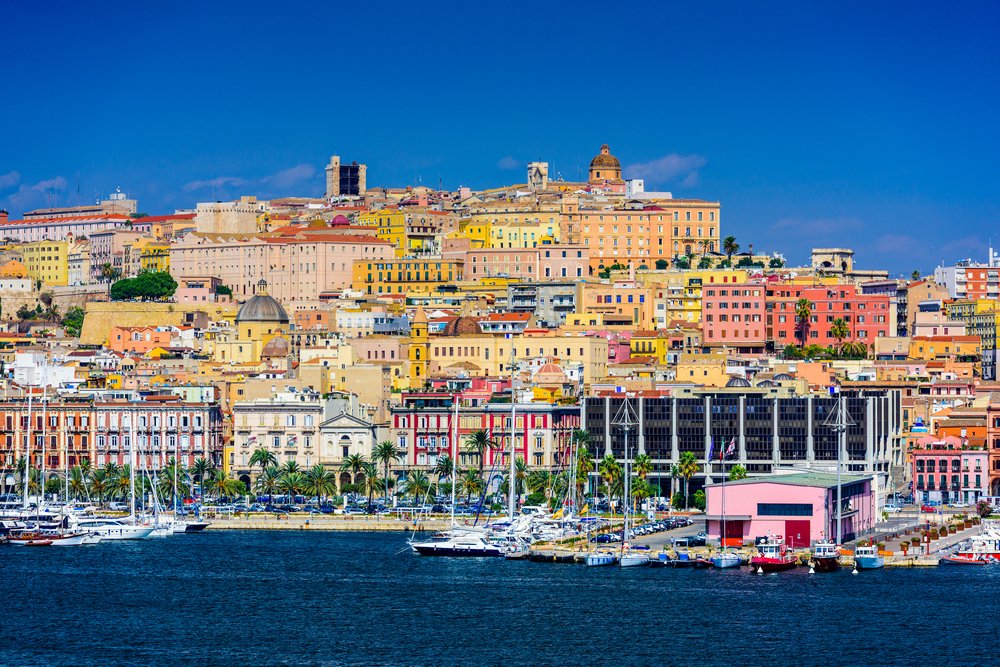 The lively capital city of Cagliari, with a marina, colorful houses in shades of yellow, red, pink, and orange all built into a hill. The sky is clear and sunny and the water appears calm.