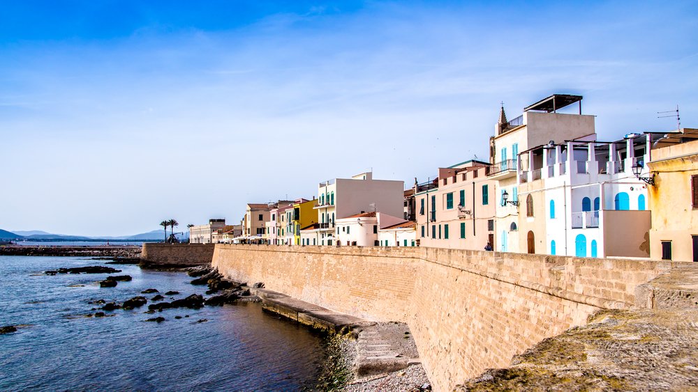 waterfront of alghero in sardinia, with colorful houses on the seaside with a sea wall and sunny sky
