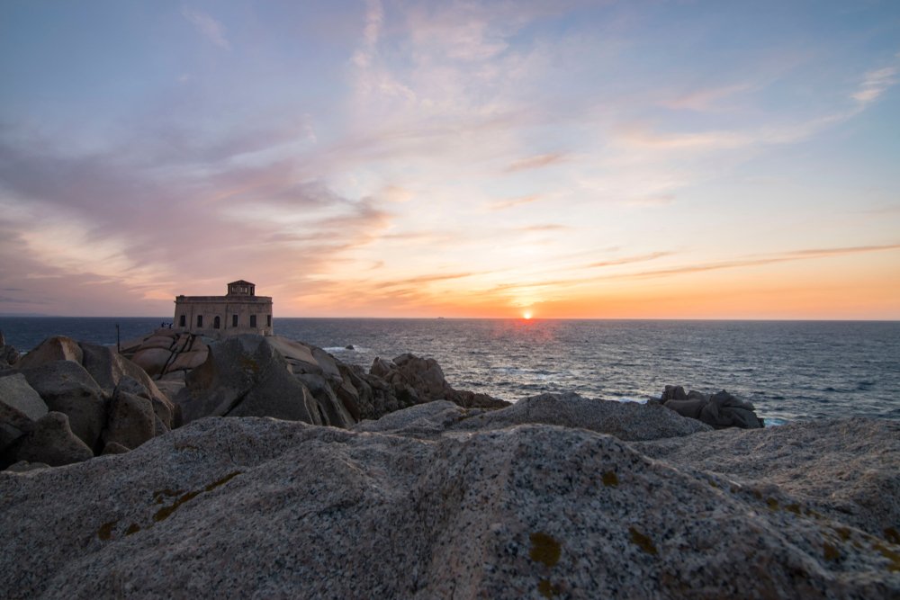 Sunset at the Capo Testa lighthouse in Sardinia, the sun sinking into the horizon with orange streaks and lighting up the clouds, the lighthouse silhouetted in the rocky foreground.
