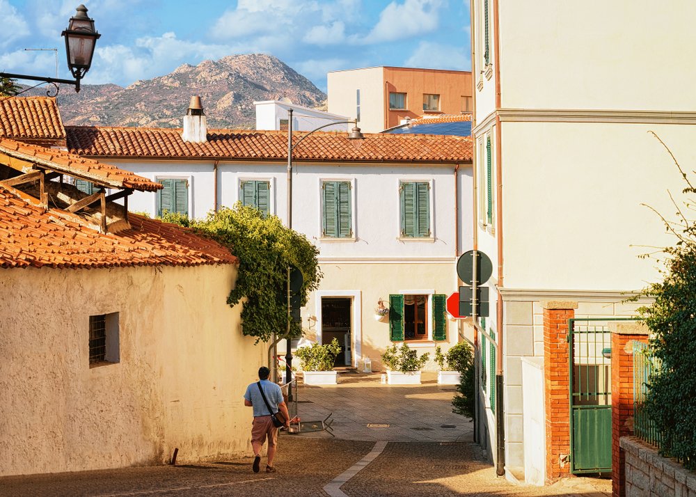 Cozy street in Olbia, Sardinia, a man walking down an otherwise empty street, with white buildings and mountains in the background