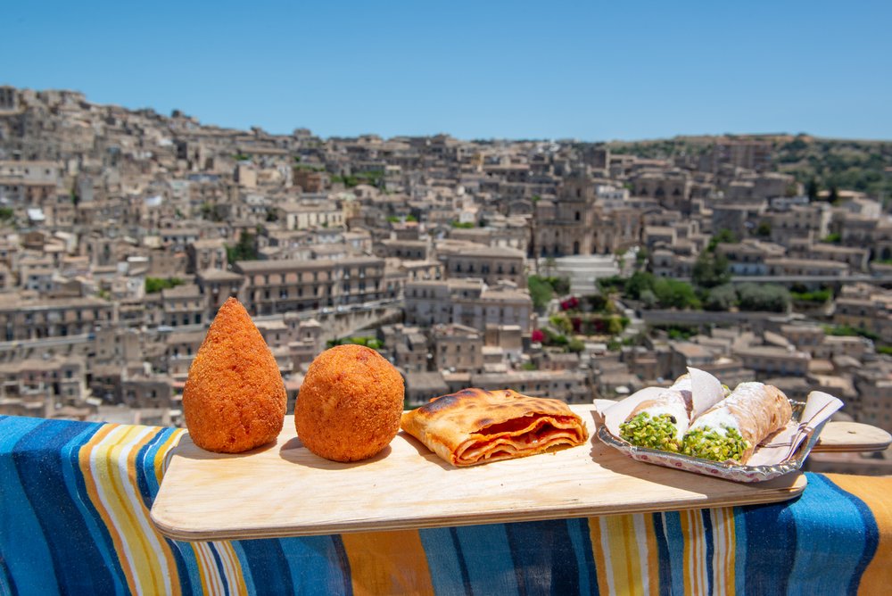 Traditional Sicilian snacks like arancini, pastry, and cannoli in front of the city of Modica, Sicily