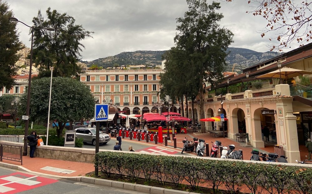 A street and square in downtown Monaco, with bike rentals, covered terrace for outdoor dining, archways and building, with mountains in the background. The sky is overcast and gray, and there are only a few people out and about and they are wearing warm clothes.