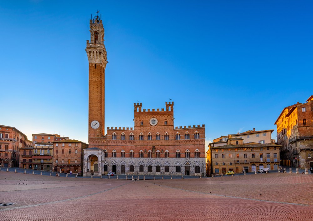 Palazzo Pubblico's gothic facade with archways, red stone, and a large campanile bell tower with a clock on it, in an empty piazza