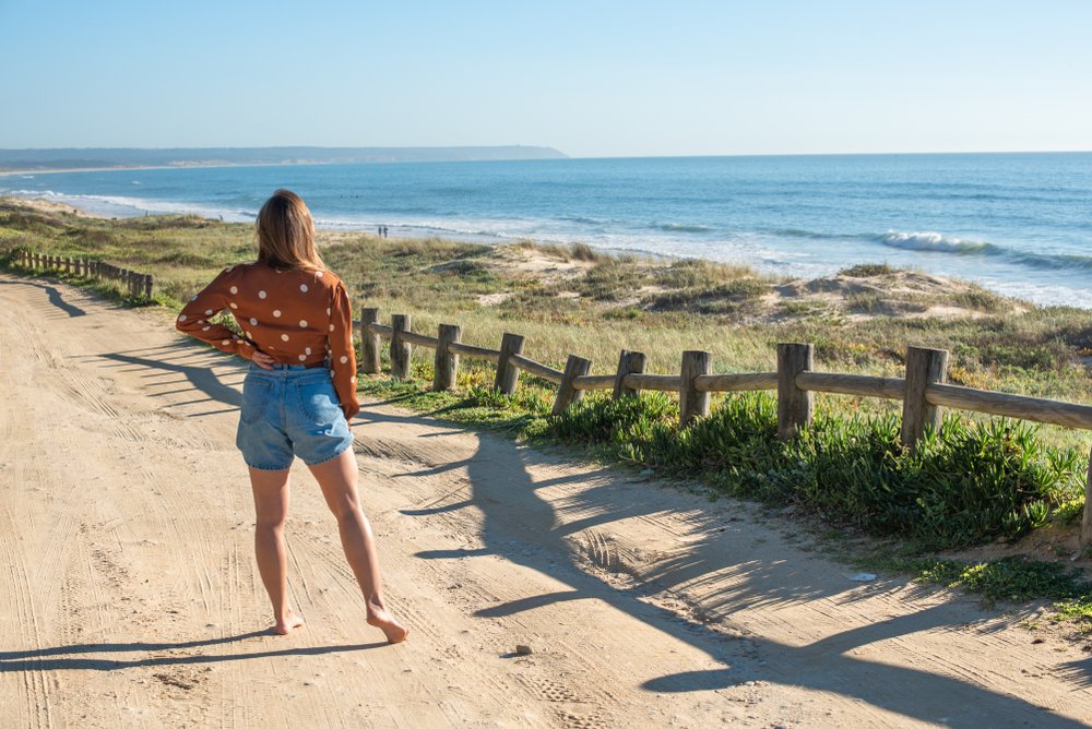 woman standing in front of the beach, with a fence between the trail and the beach, looking out onto the view