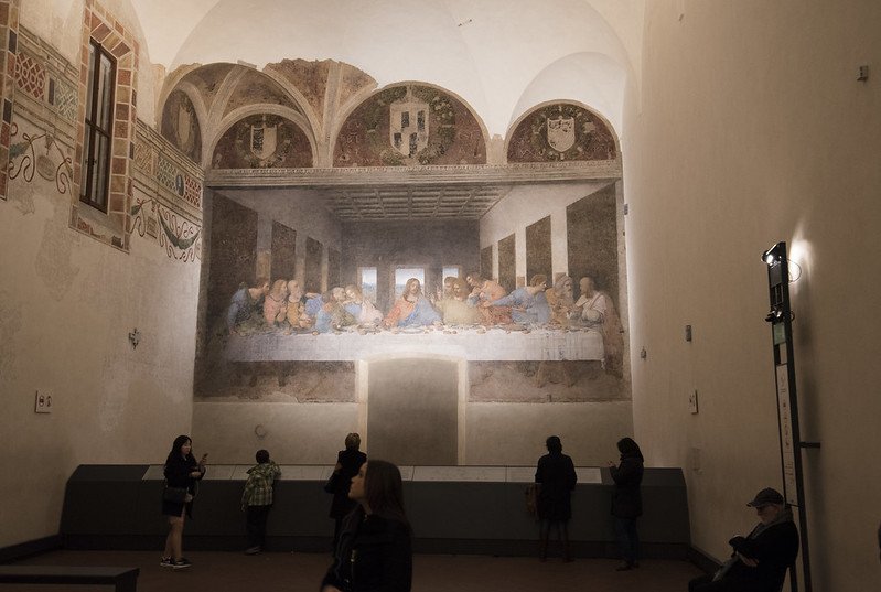 a handful of people inside the last supper room of the church, looking at the fresco, with small lights and guard