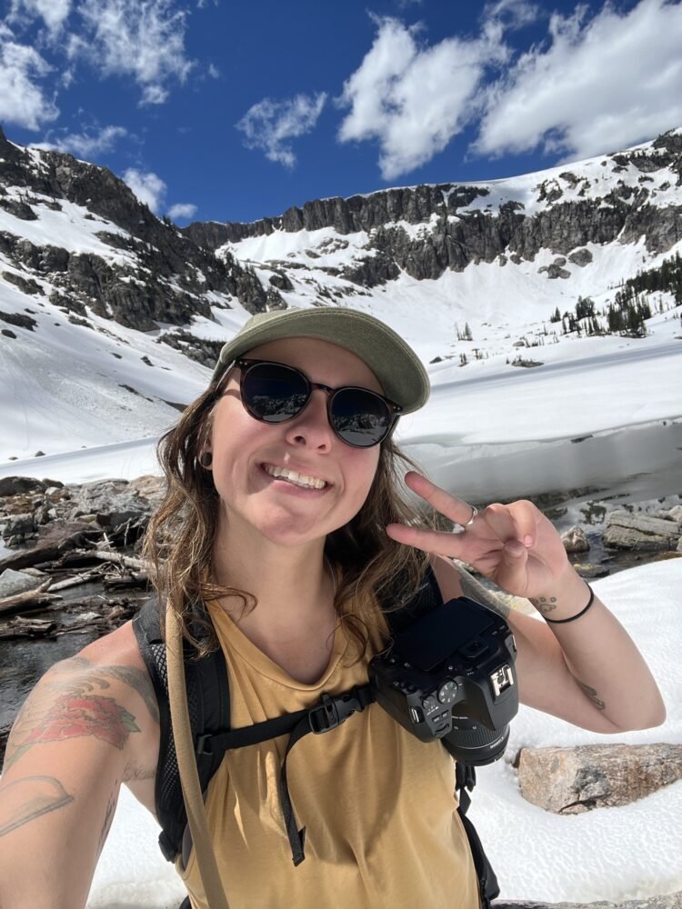 The author, Nicole Westcott, visiting one of the many lakes of Grand Teton National Park in a snowy part of the park wearing a yellow tank top on a summery day