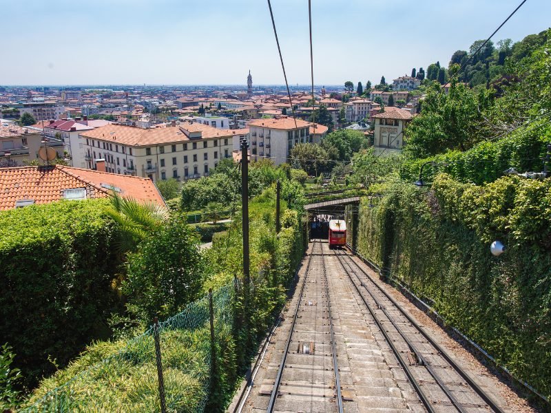 A very steeply graded railway with a red train car working its way up the track, with views of the city of Como below it.