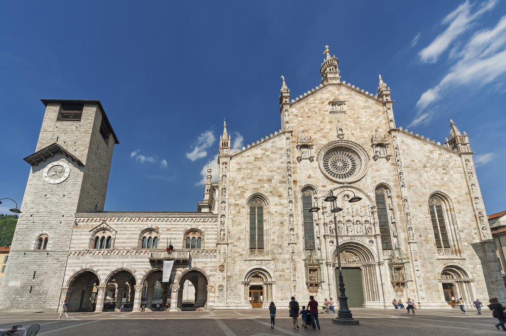 Beige stone facade of Cathedral in Como city (also known as the Duomo) with a church building, archways, and a belltower. Some people in front of the church during the middle of the day.

