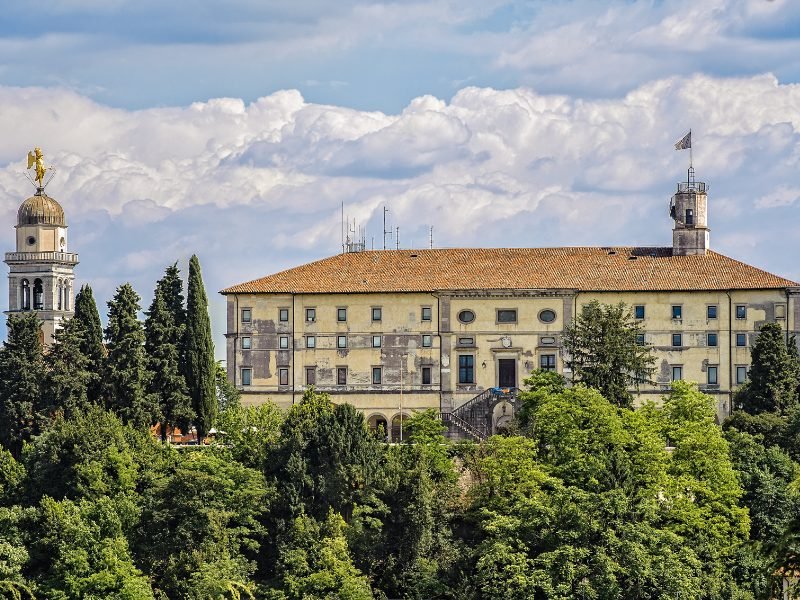 The Udine Castle with treetops covering up the wall leading up to the castle, with a bellfry visible, and the castle is yellowish with peeling paint and flying a flag on top.