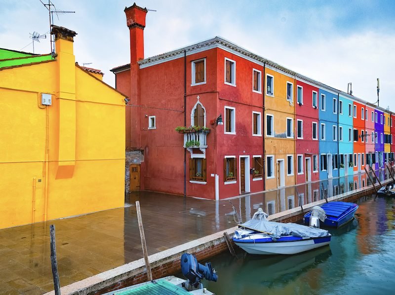 Yellow House, red and other color houses brightly painted on a canal with some small boats on an island in the outskirts of Venice