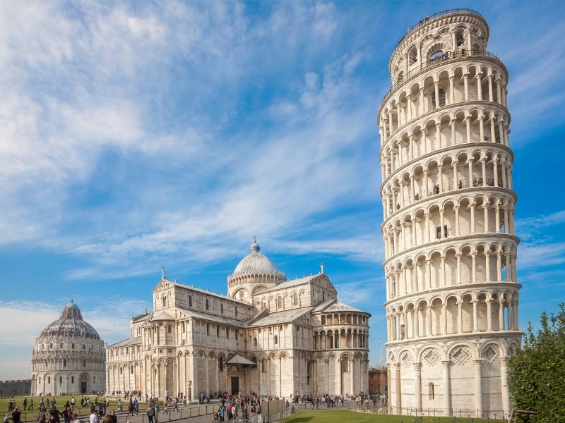 The many-arched ornate leaning tower of pisa, towering over the cathedral and offering an optical illusion, on a sunny day in the summer with lots of tourists out and about enjoying the unesco site of pisa.