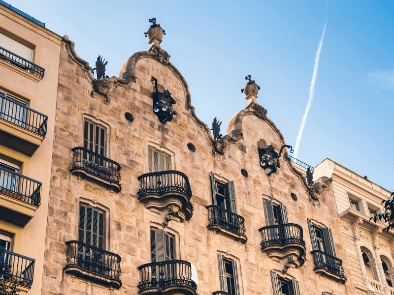 View of the facade of Casa Calvet, a lesser-known Gaudi building, with orange balconies that bulge from the facade of the building, on a sunny day with an airplane trail in the sky