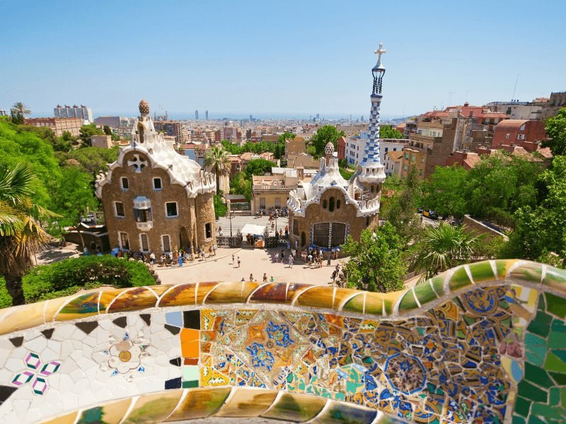 View of the balcony of Park Guell with lots of mosaics and building colors and gingerbread-looking architecture with funky towers, view of the city of Barcelona below it and even down to the beaches