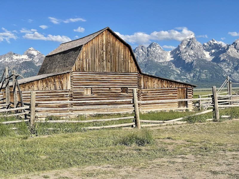 Mormon Row in Grand Teton National Park with an old farmhouse and mountains behind it on a sunny day with just a few clouds in the sky