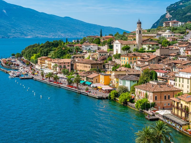 Clear waters of Lake Garda, with the town of Limone Sul Garda going up the hillside, and a lakefront promenade where people enjoy a walk