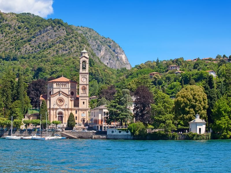 historic pinkish beige church with belltower on the lake with trees and mountains behind it.