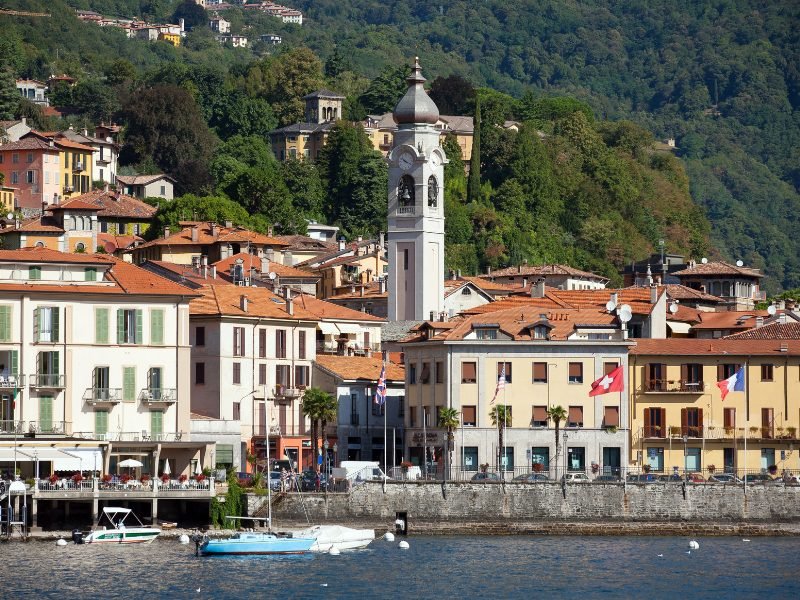 The waterfront town of Menaggio with a tower with a clock and a bunch of flags along the waterfront and hills in the background