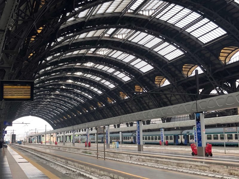 The Milan train station with its rounded open-window style train station with a train on the faraway track and an electronic train schedule