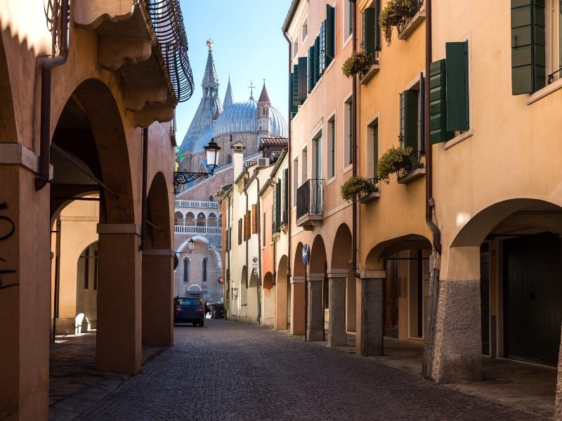 Views in the city of Padua, Italy, with porticos and archways and a church in the background at the end of the narrow street, one car in the street.