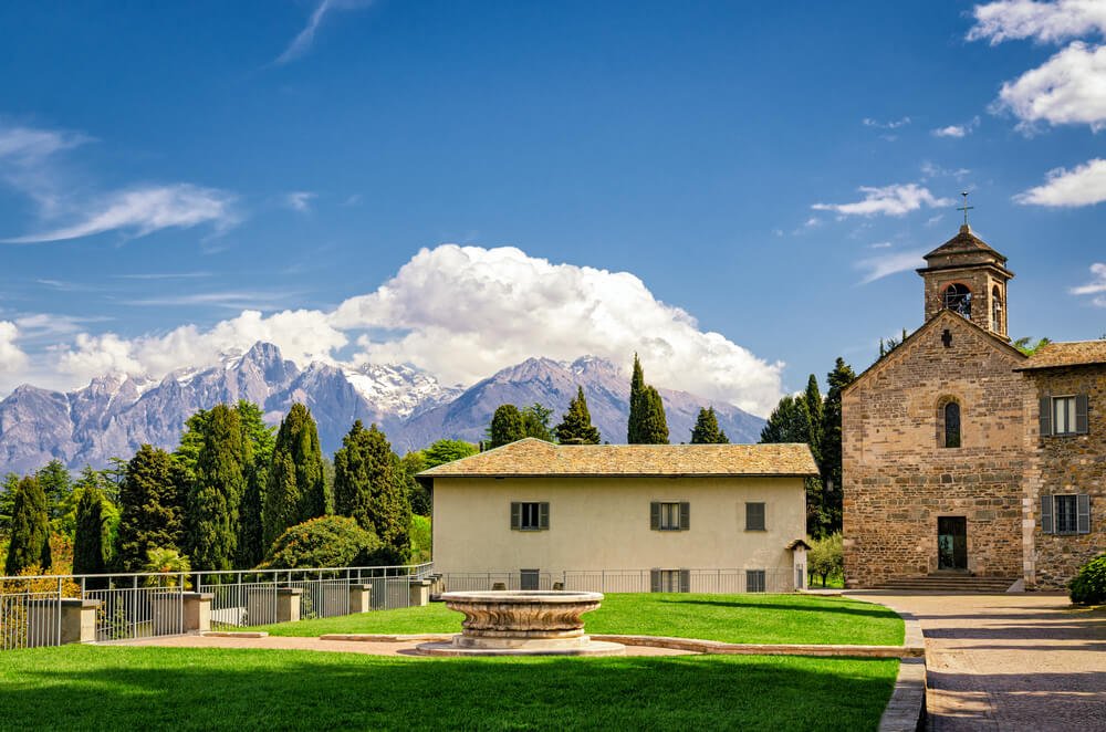 The terra-cotta colored brick building that is Piona Abbey in the  Lake Como area with views of high mountain peaks in the distance, and evergreen trees.