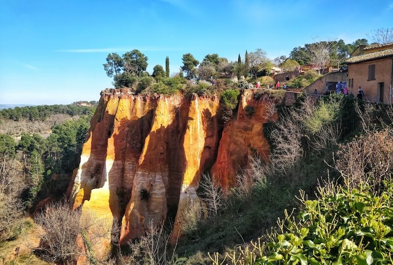 the ochre red and orange cliffs of the town of roussillon in provence's luberon valley, with other brush and plant life, as late afternoon light falls on the cliff
