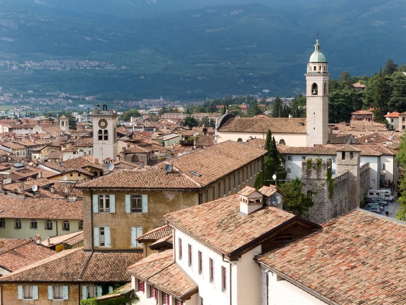 Bell tower, church towers, and buildings as you admire the landscape of Rovereto, with foothills in the background.