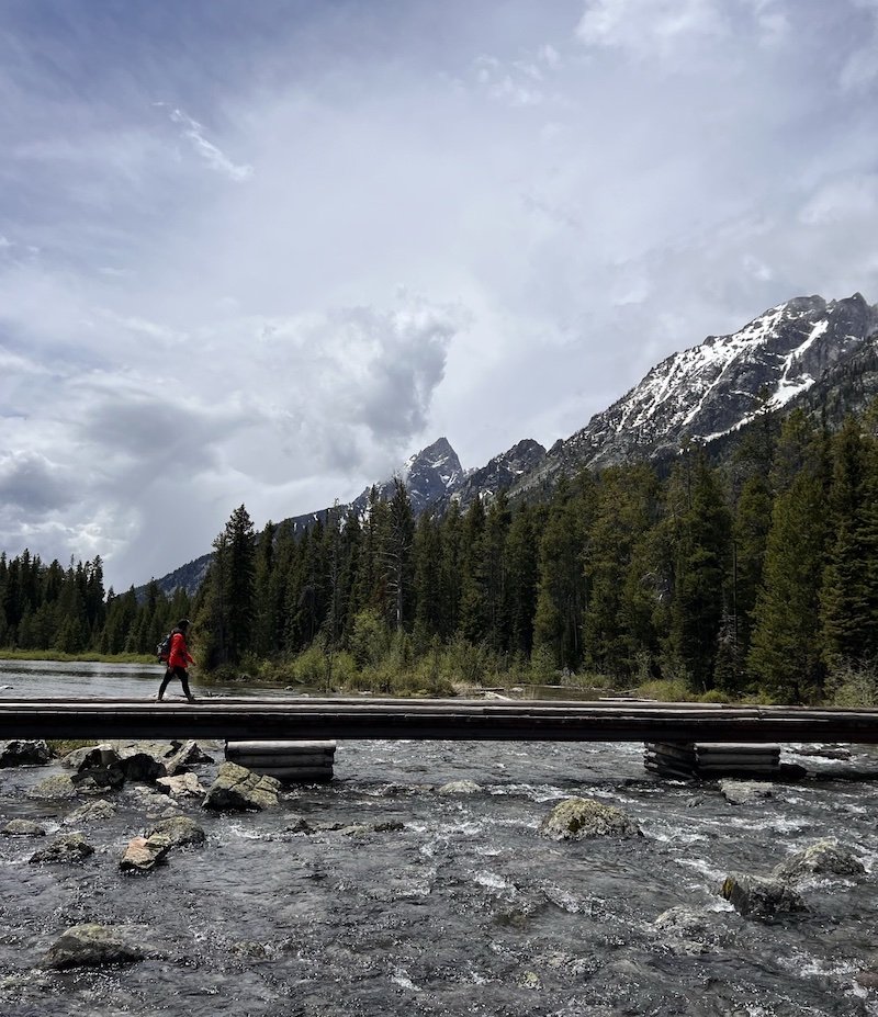 Nicole Westcott hiking on the path towards String Lake, crossing a pedestrian bridge over a river, towards the Teton Range