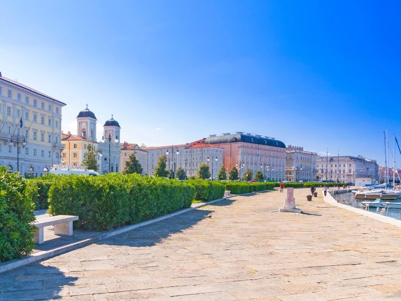 The port city of Trieste in Northern Italy, walking along the waterfront with boats and a waterfront promenade, buildings behind it.