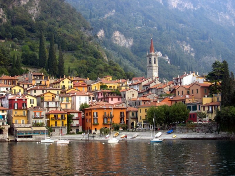 The town of Varenna as seen from the water with orange, yellow, and other colorful houses on the lakefront, and a large clock tower with red steeple, and mountains behind it.