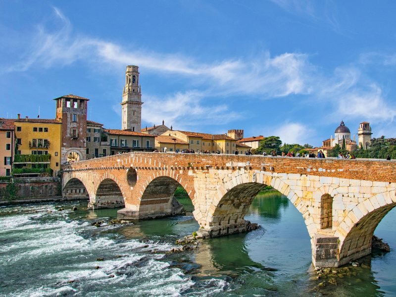 People on a very old historic bridge that crosses the river to the main town center of Verona, with historic buildings and churches