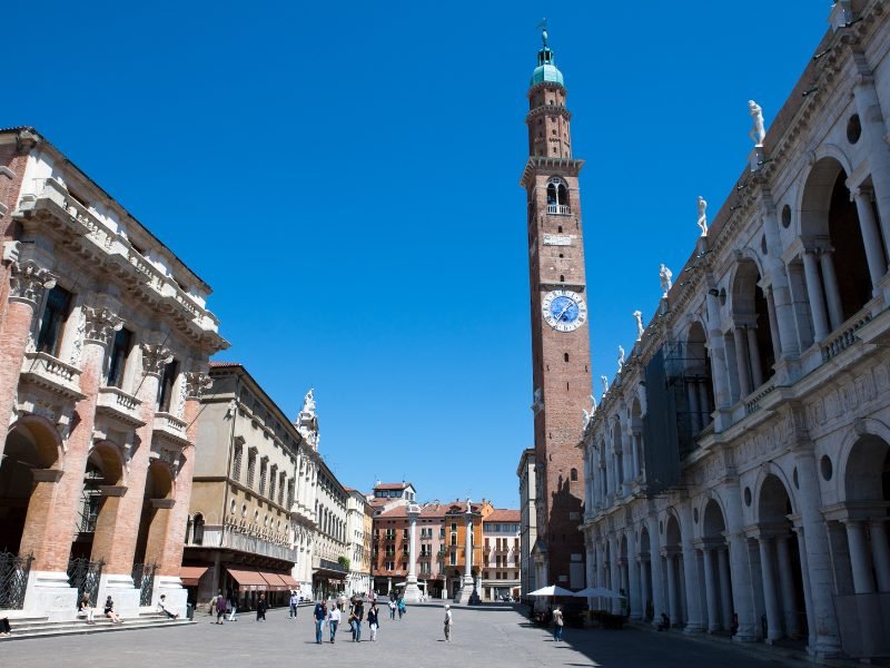 A large tower in Vicenza with a clock on it, also a piazza and people walking in the square, old-fashioned buildings on a sunny day