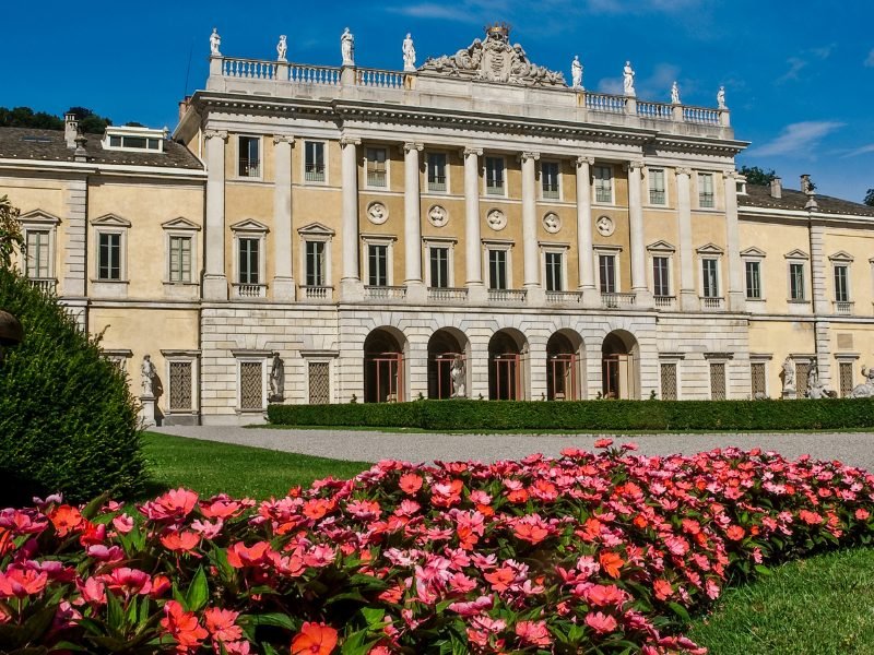 Yellow facade of Villa Olmo on the outskirts of Lake Como with red flowers in the springtime in front of the villa and grassy lawn.