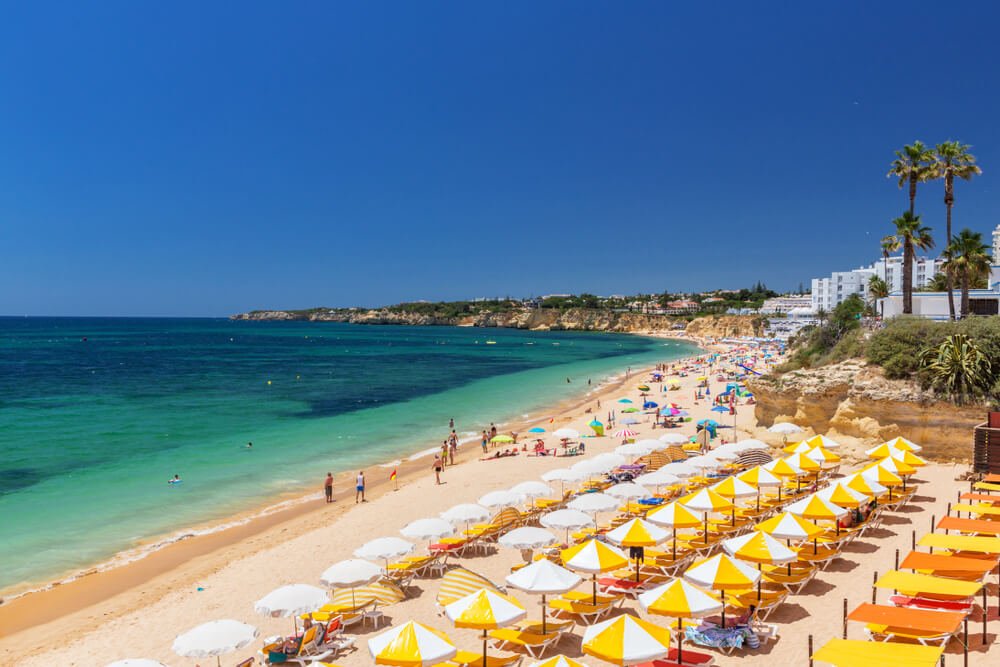 Beautiful beaches of the Algarve coast of Portugal, Armacao de Pera, with brilliant yellow and white umbrellas, turquoise and dark blue water, soft white sand, and people enjoying the beach on a summer day.