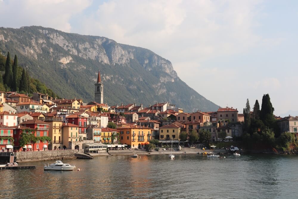 View on Bellano at Lago di Como, with a small white boat in front of the waterfront, red and yellow buildings with one tall belltower for the local church, with mountains towering behind it