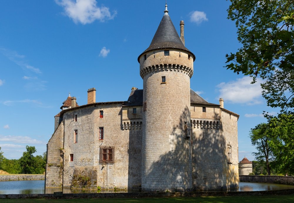 big circular turret and a castle, with a lake or moat or some sort of water behind it, in nature with lots of trees and grass