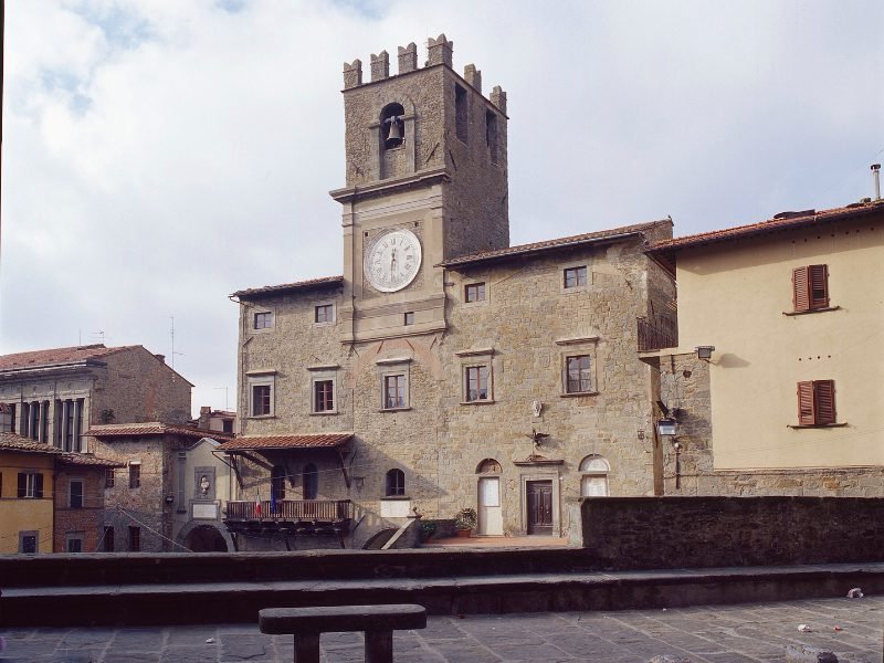 the palazzo communale of cortona, with medieval clock tower with rectangular pillar, benches for pedestrians on a cloudy day.