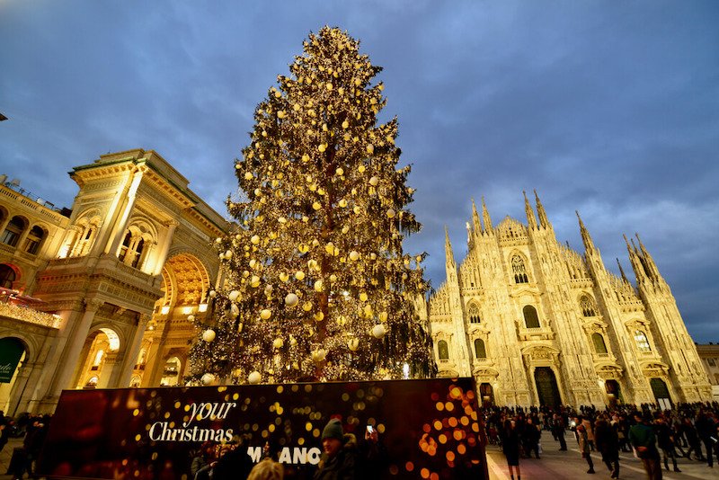 The christmas tree draped in gold lights and ornaments in front of the milan duomo and lots of people milling about the facade of the duomo visiting market stalls in milan at christmas