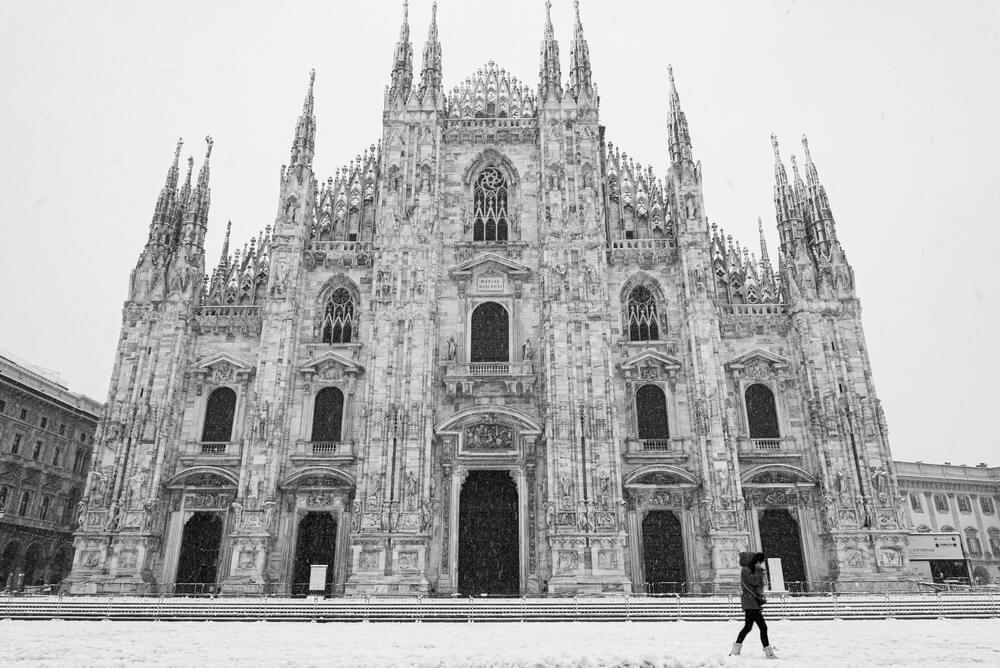 A rare snowy day in Milan in winter with snow on the ground and a person in snow boots and a winter jacket walking in front of the marble facade of the Milan Duomo