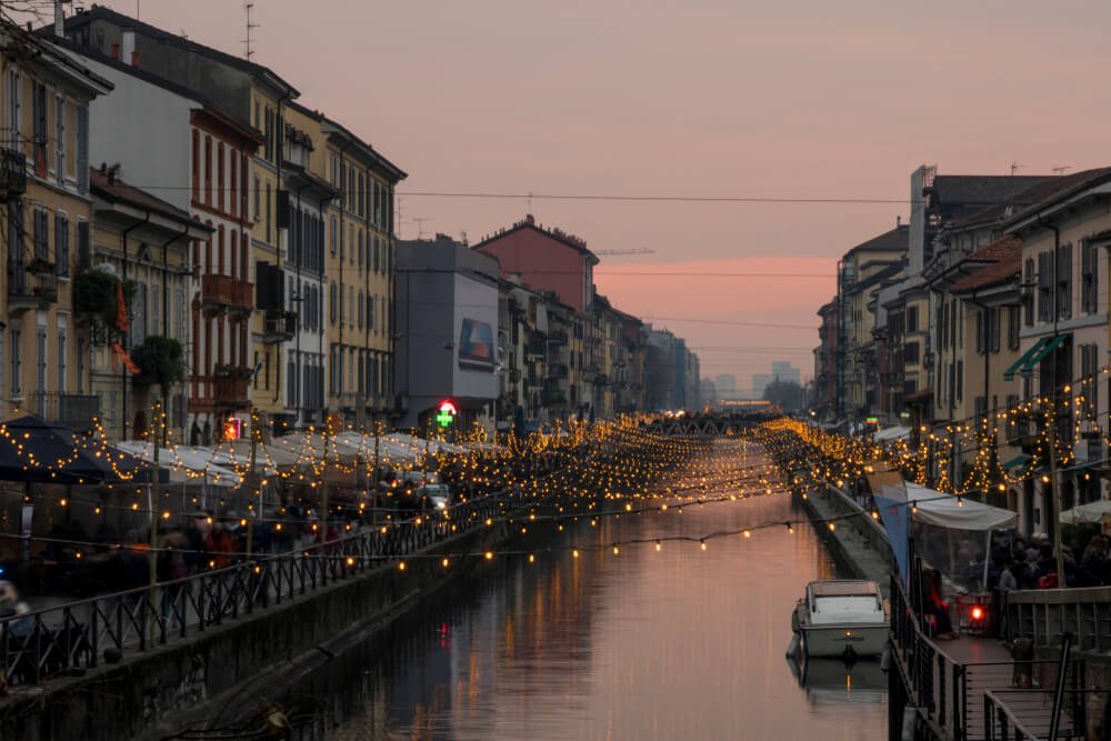 Milan at Christmas with lights on the Naviglio Grande canal waterway at evening: Navigli is a district is famous for its restaurants, cafes, pubs and nightlife. Night sky is pink and purple and lights are golden.