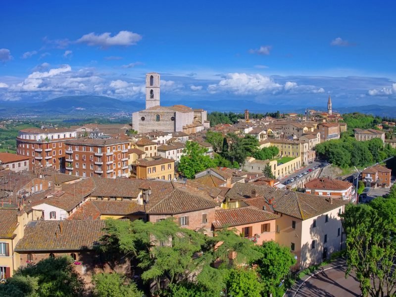 The hilly town of Perugia in Umbria with lots of buildings, several churches with steeples and towers visible, and outer hills