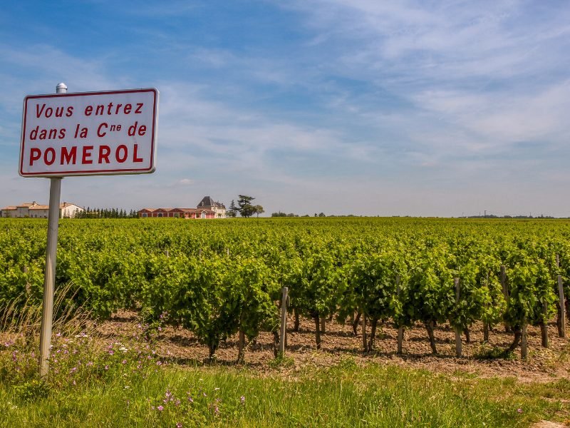 sign that says, translated from the french, 'you are entering in the commune of pomerol' with vineyards and some agricultural buildings like barns, farmhouses in the background